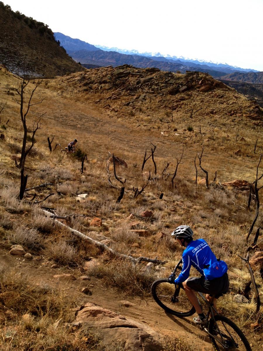 A mountain biker in a blue jersey navigates a rocky trail on a rugged hillside, surrounded by dry grass and charred tree remnants, with distant mountains visible in the background under a clear sky. Oil Well Flats mountain bike trail.