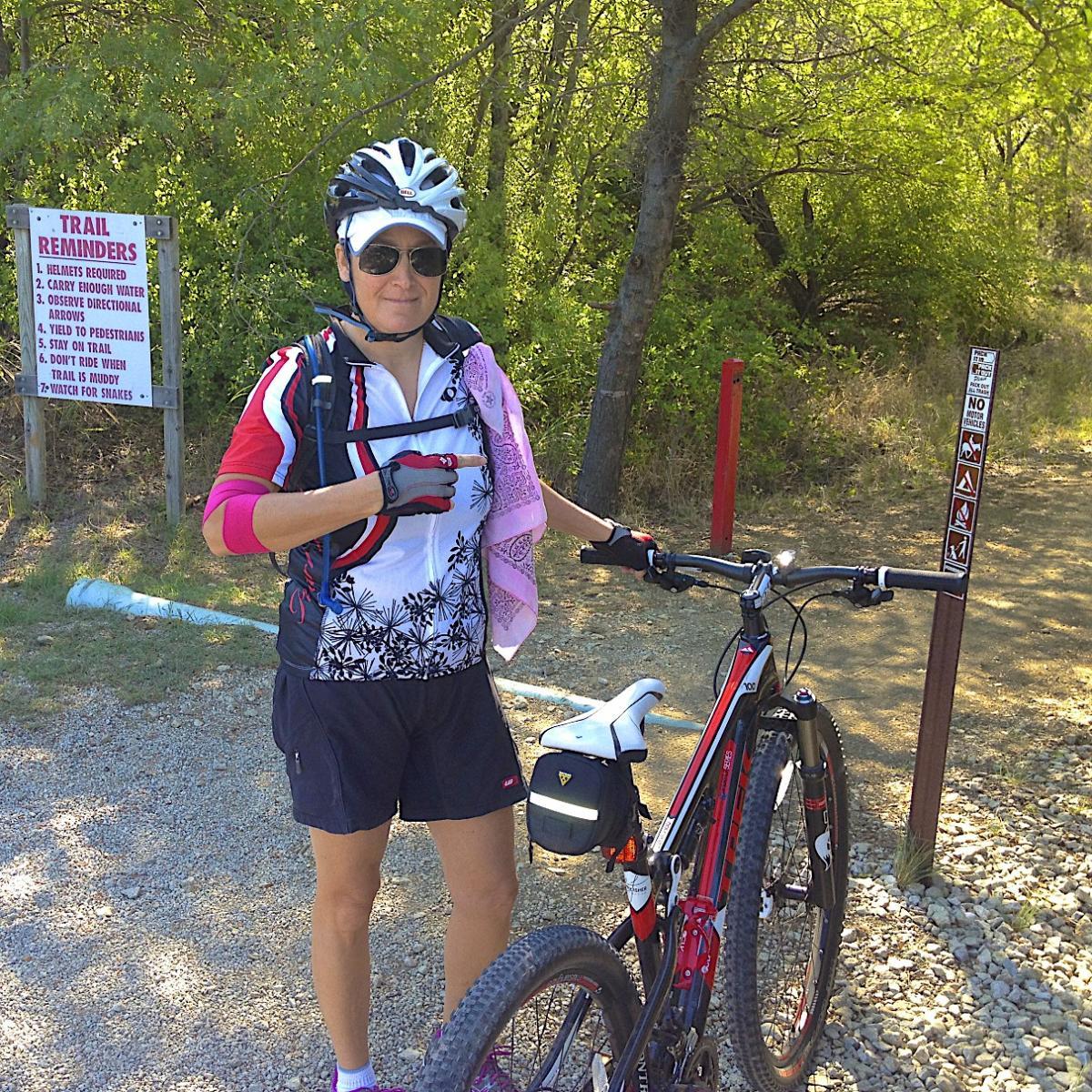 A cyclist stands next to a mountain bike, wearing a helmet and sunglasses. She is dressed in a colorful cycling jersey and shorts, with a headband and a towel draped around her neck. In the background, there is a sign with trail reminders and regulations, surrounded by greenery and a gravel path. Cedar Hill State Park At Joe Pool Lake mountain bike trail.
