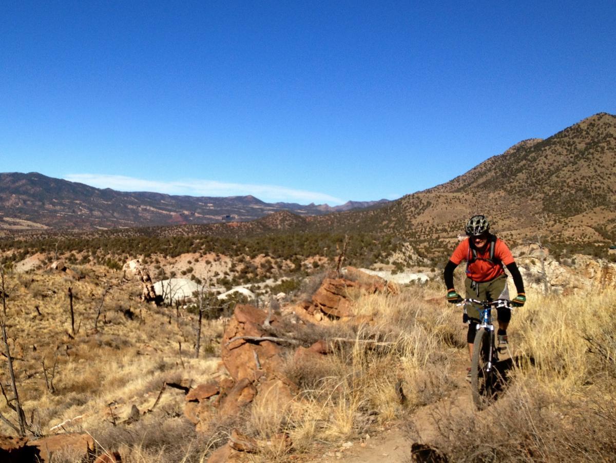 A mountain biker navigating a rocky trail in a rugged landscape, surrounded by distant mountains and a clear blue sky. The terrain features dry grass and scattered rocks, showcasing a scenic outdoor adventure. Oil Well Flats mountain bike trail.