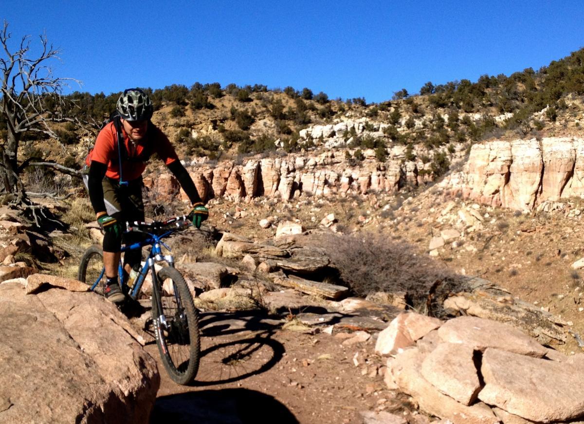 A mountain biker maneuvering over rocky terrain on a sunny day, with rugged cliffs and sparse vegetation in the background. The cyclist is wearing a helmet, gloves, and an orange shirt, showcasing an adventurous outdoor activity. Oil Well Flats mountain bike trail.