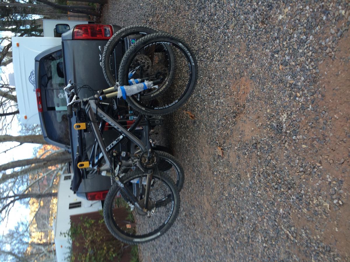 Trek Remedy 7: A mountain bike mounted on a rack attached to the back of a pickup truck, parked on a gravel driveway surrounded by trees.