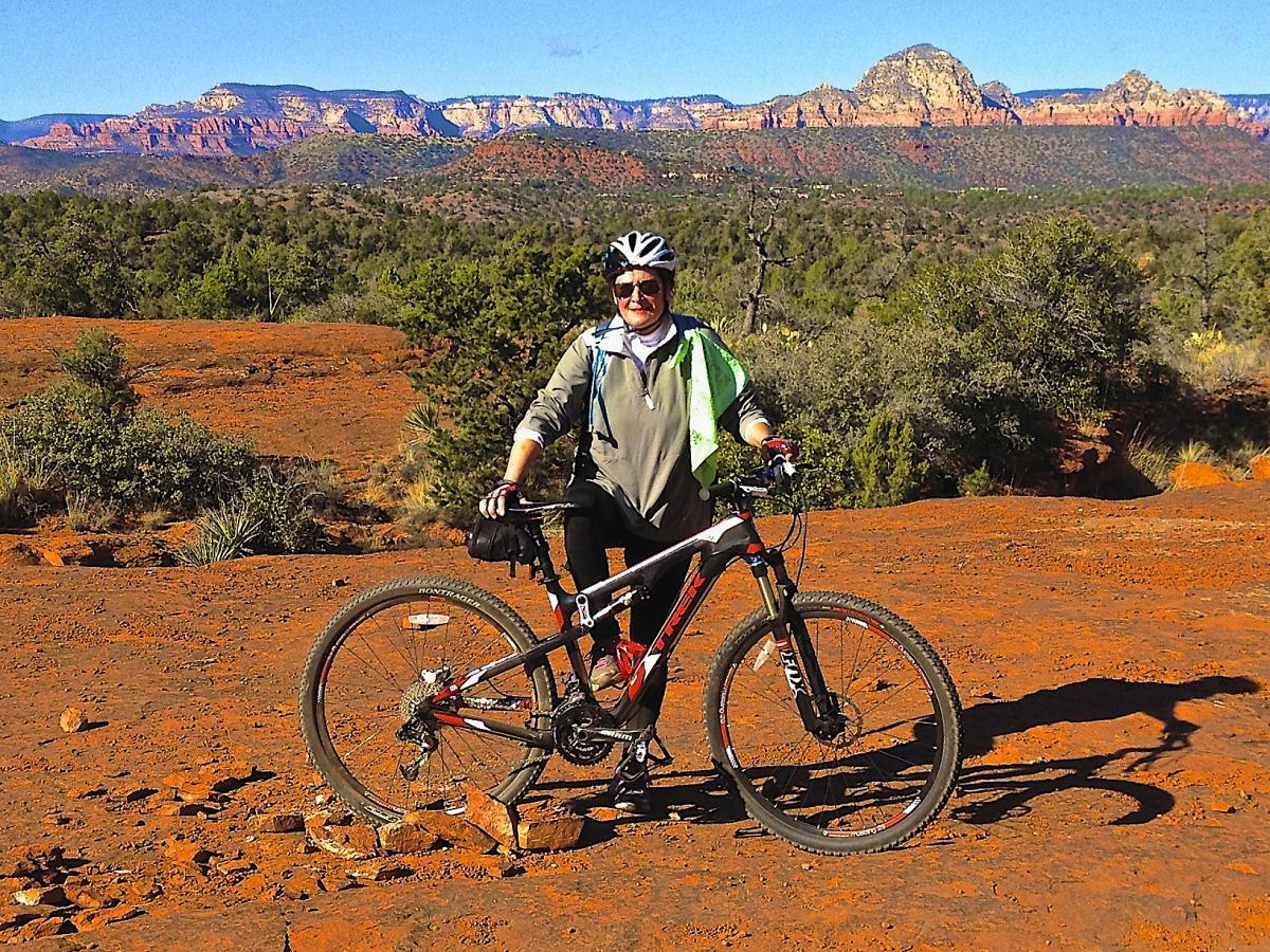 A person standing next to a mountain bike on a rocky, red landscape with green shrubs and distant mountains in the background under a clear blue sky. The individual is wearing a helmet, a light jacket, and has a towel draped over their shoulders, smiling at the camera. Bell Rock Trailway mountain bike trail.