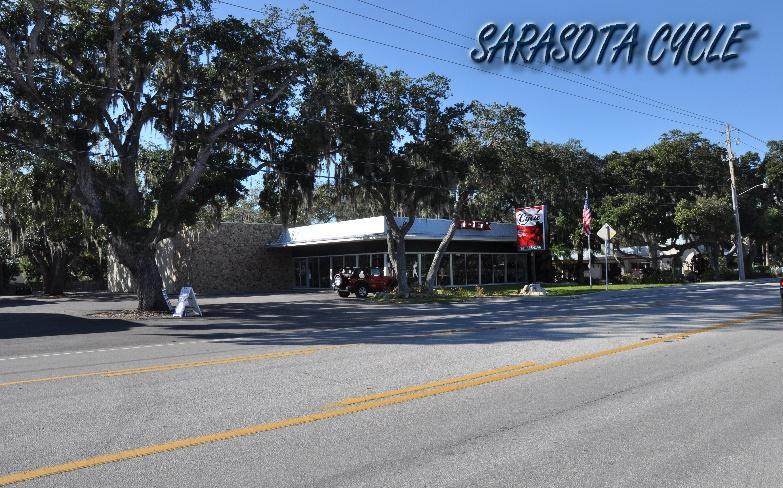 A roadside view of Sarasota Cycle, a bicycle shop, featuring a modern building with large glass windows. The shop is surrounded by lush trees with Spanish moss, and there are flags visible in the vicinity. The image captures the storefront from across the street, showcasing the inviting atmosphere of the area. The text "SARASOTA CYCLE" is displayed prominently in the corner.