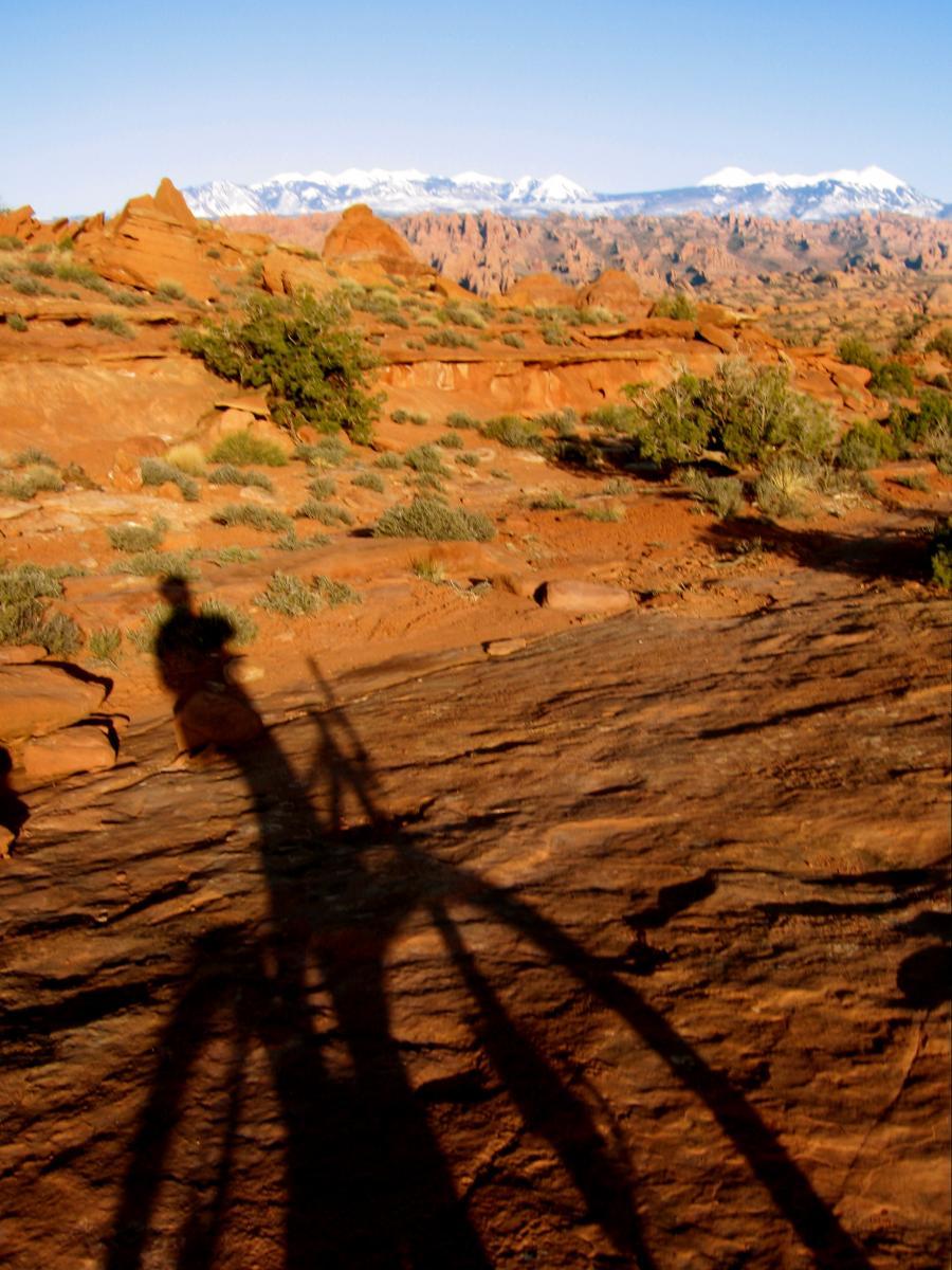 A silhouette of a person and their bicycle cast on rocky terrain, with a backdrop of snow-capped mountains and clear blue sky. The scene is set in a desert landscape, featuring red rocks and sparse vegetation. Captain Ahab mountain bike trail.