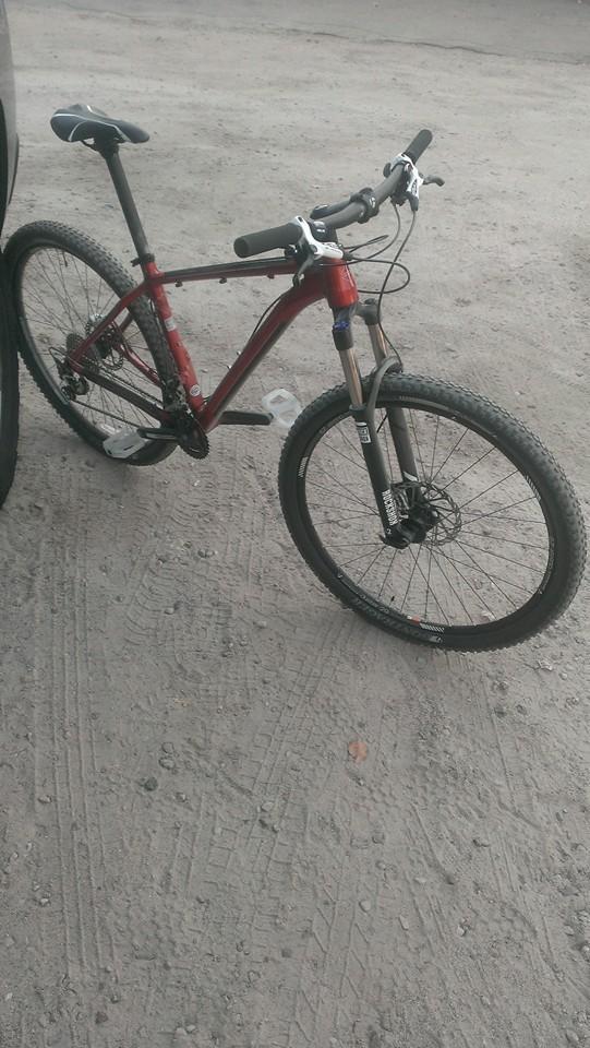 Trek Stache: A red mountain bike with black tires and front suspension is positioned on a dirt path, partially leaning against a vehicle. The bike's handlebars are visible, featuring black grips and a small water bottle attached to the frame. The surrounding area shows textured dirt with faint tire tracks.