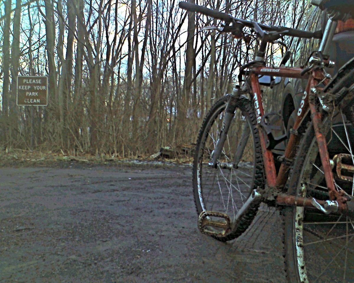 Trek 820: A close-up view of a mountain bike with muddy tires parked on a gravel surface, with a background of bare trees. A sign in the background reads, "PLEASE KEEP YOUR PARK CLEAN." The scene is set in a natural area, suggesting a focus on outdoor activities.