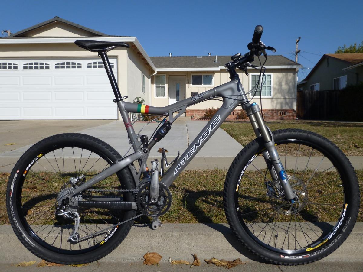 A silver mountain bike parked on a sidewalk in front of a residential house. The bike features wide tires, a dual suspension system, and colorful accents on the seat post. In the background, a single-story home with a garage and landscaped yard is visible under a clear blue sky.