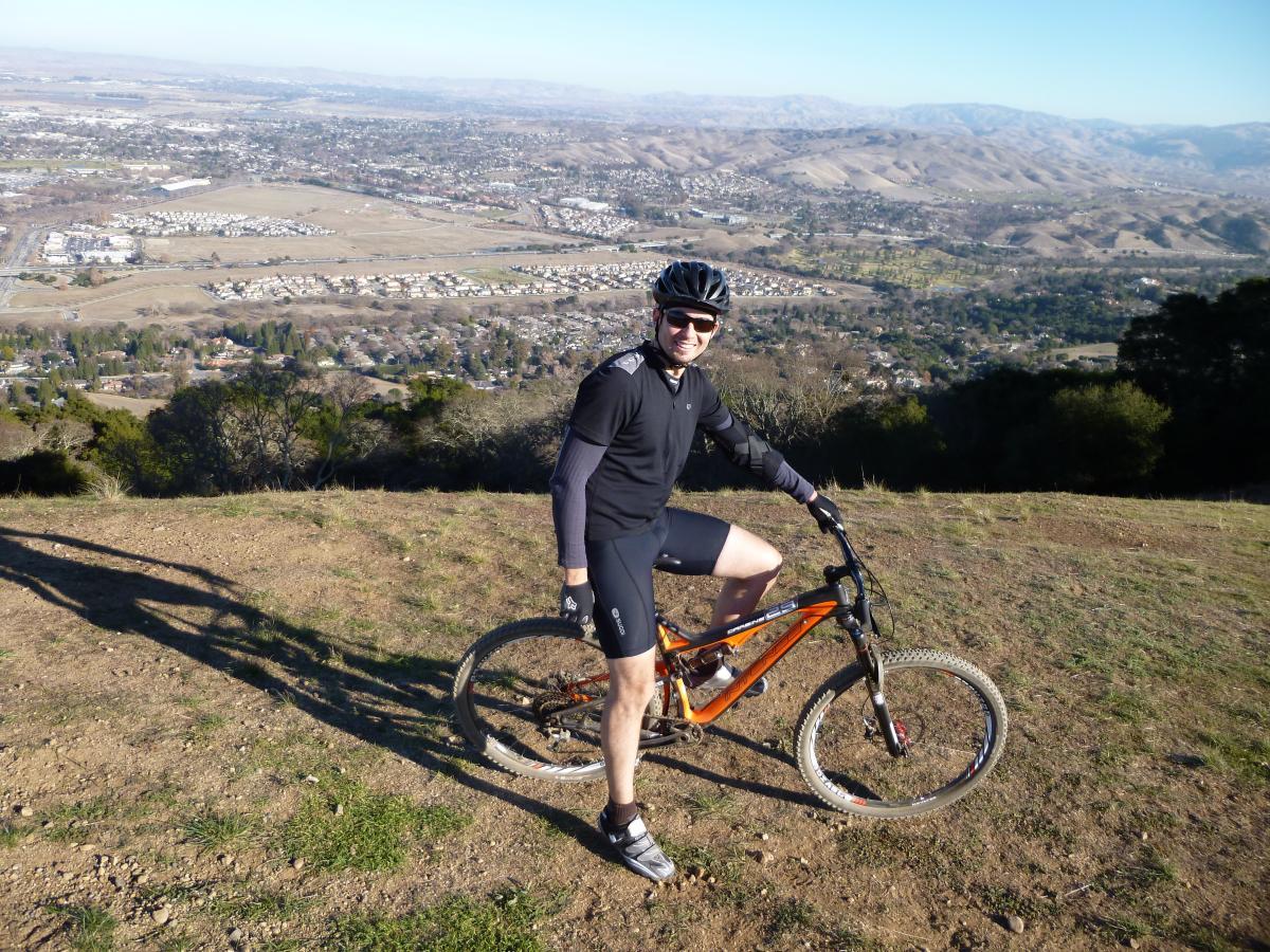 Intense Carbine 29: A cyclist poses on a mountain bike at the edge of a hill, overlooking a scenic valley with rolling hills and residential areas in the background. The cyclist is wearing a helmet, sunglasses, and athletic clothing, and the sun casts a shadow on the grassy terrain.