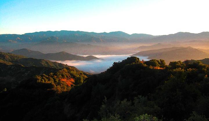 A panoramic view of rolling hills and mountains bathed in soft morning light, with a layer of fog gently resting in the valleys below. The landscape features lush greenery on the foreground hills, while distant peaks are silhouetted against a clear sky, creating a serene and tranquil atmosphere. Tour De Los Padres mountain bike trail.