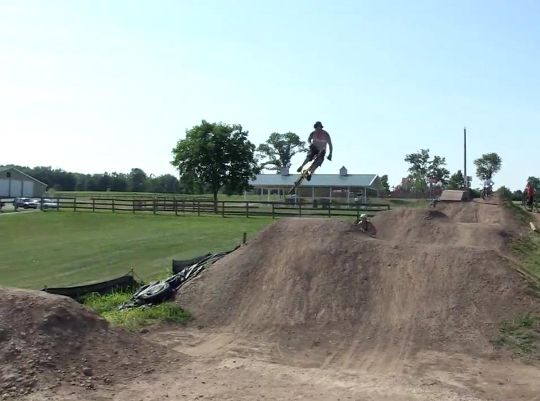 A person performing a jump on a BMX bike over a dirt mound at a biking park, with a grassy area and trees in the background. A house and other riders can be seen in the distance. The sky is clear and bright. Alexandria Pump Track mountain bike trail.