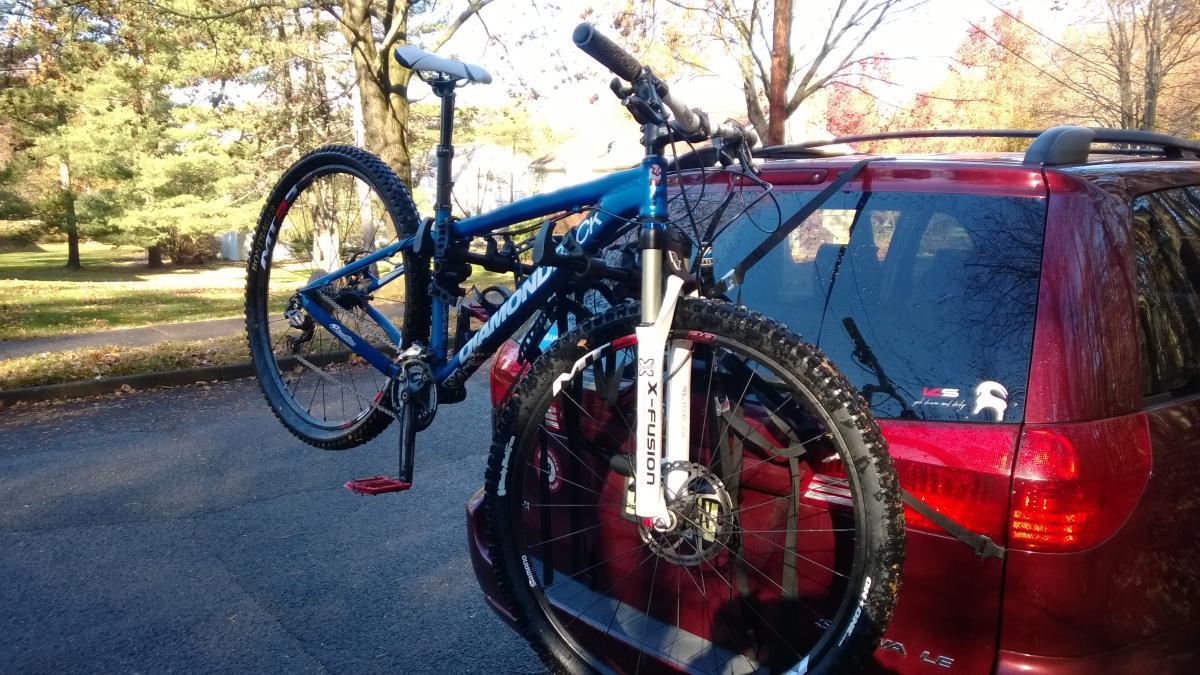 Diamondback Overdrive Sport: A blue mountain bike is securely mounted on the back of a red car, with trees and an autumn landscape visible in the background. The bike features visible tires and a suspension fork, and is attached using bike racks seen in the image.