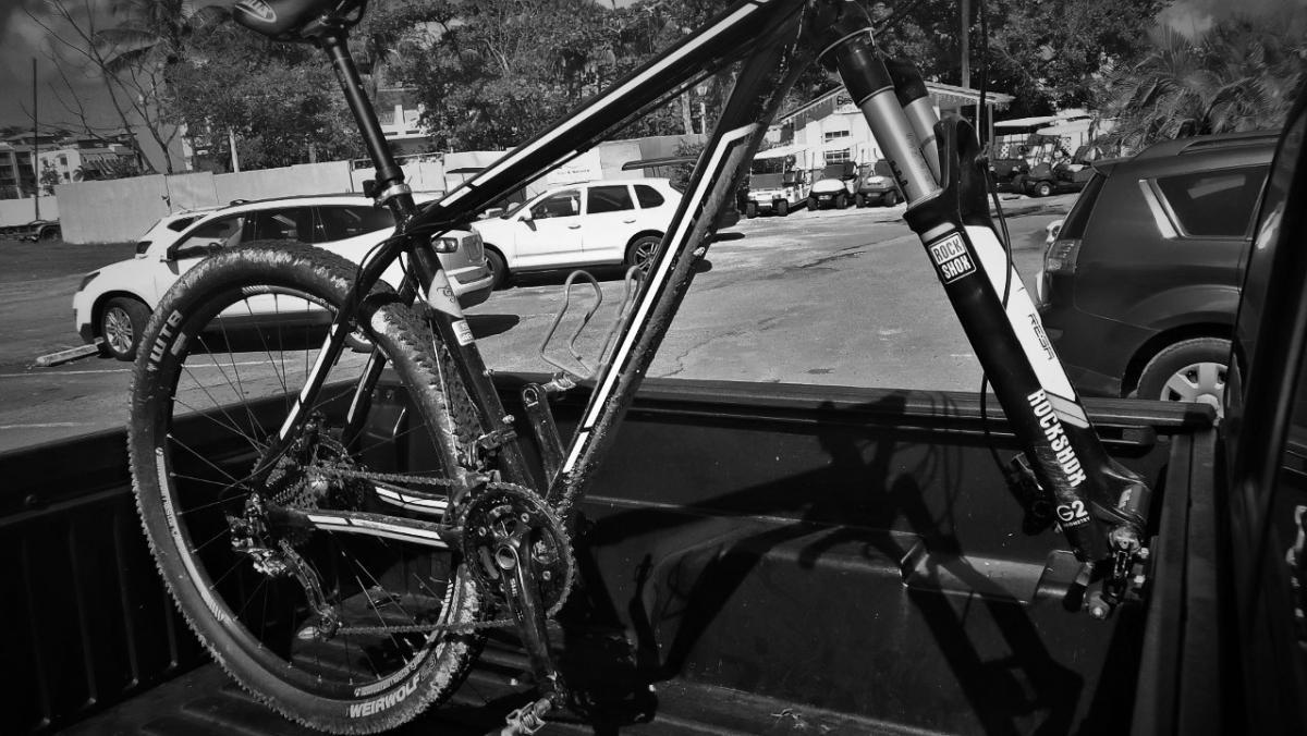 Trek Superfly AL: A mountain bike secured in the back of a pickup truck, showcasing its frame, wheels with dirt markings, and a visible shock absorber. The background includes parked vehicles and trees, captured in black and white.