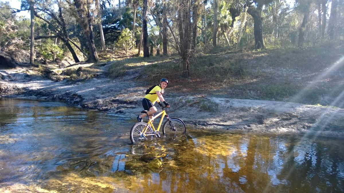 A person riding a yellow mountain bike through shallow water in a wooded area, surrounded by trees and sunlight filtering through the leaves. The rider is smiling and wearing a yellow shirt and helmet. Little Big Econ State Forest mountain bike trail.