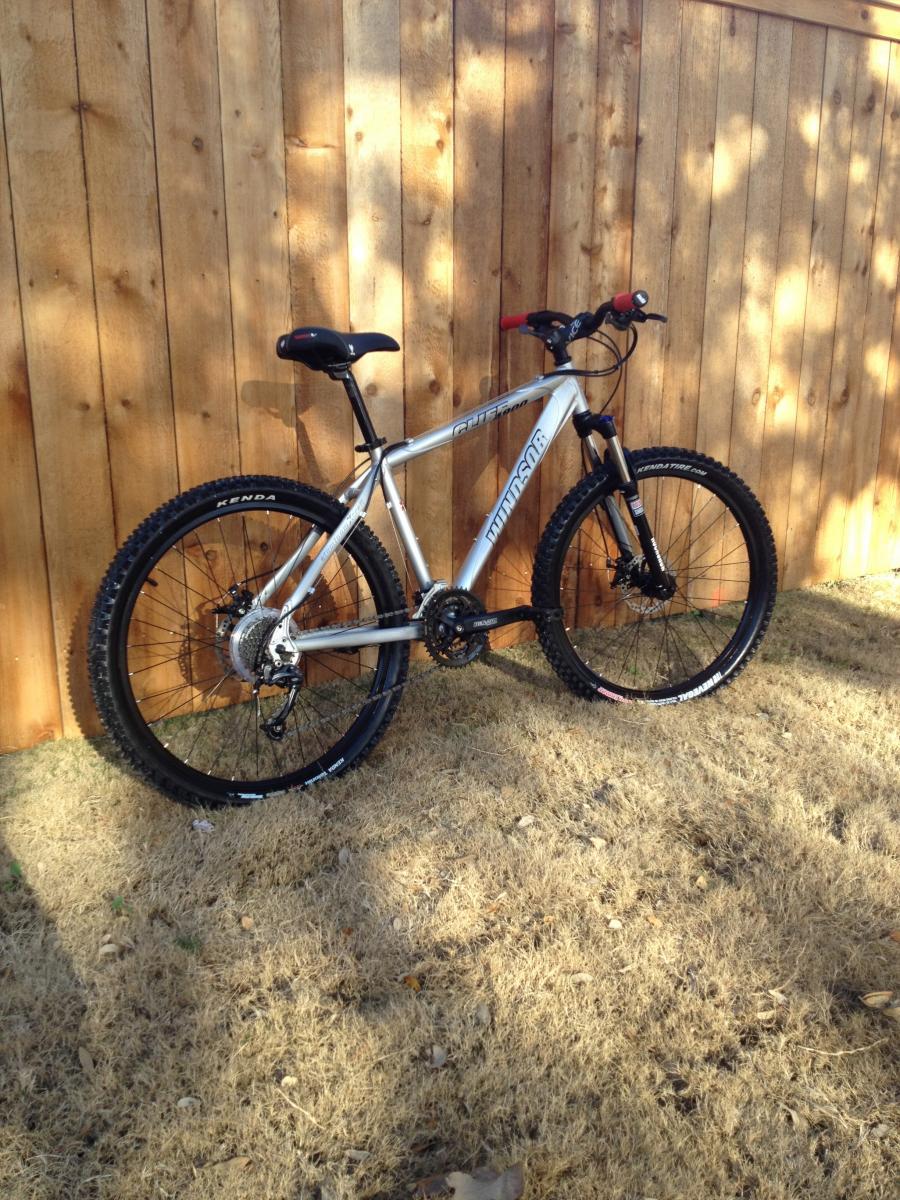 Windsor Cliff 4900: A silver mountain bike resting against a wooden fence, positioned on dry grass. The bike features thick, knobby tires and a sturdy frame, with a visible gear system and handlebars. Sunlight casts shadows on the grass, highlighting the bike's sleek design.