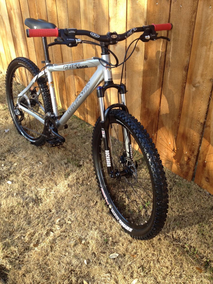 Windsor Cliff 4900: A mountain bike with a silver frame, red handle grips, and black tires, leaning against a wooden fence, with grass and scattered leaves in the foreground.