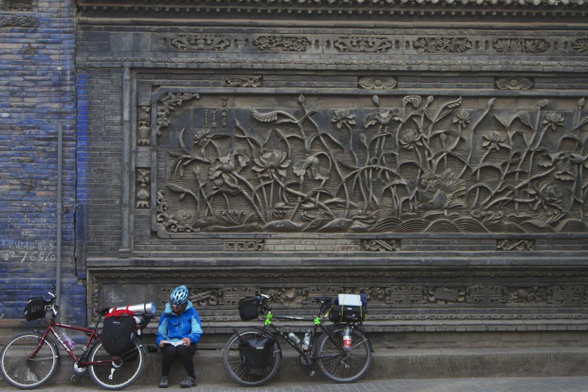 Marin Bobcat Trail: A cyclist wearing a blue jacket and helmet sits reading a book on a bench against a decorative stone wall featuring intricate carvings of flowers and plants. Two bicycles are parked beside the bench, each equipped with bags. The background wall is partially blue and showcases detailed relief artwork.