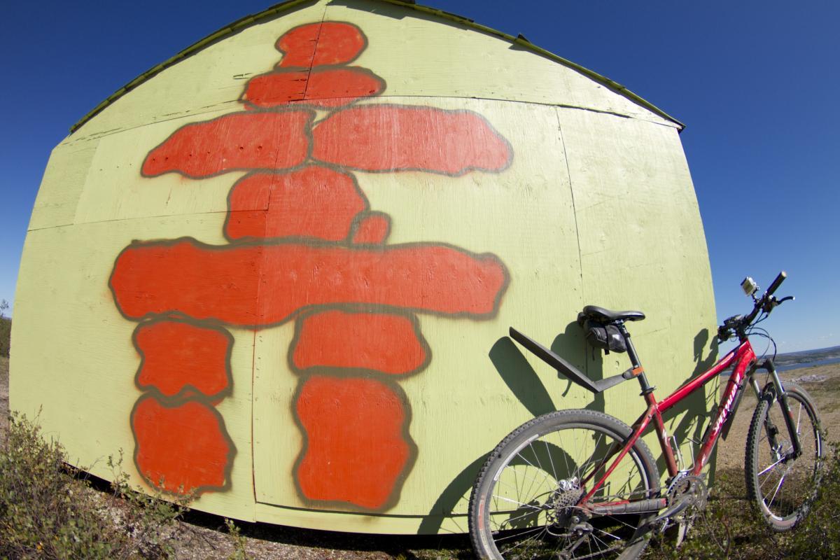 A red inukshuk symbol is painted on the side of a green wooden shed, with a mountain bike leaning against the wall. The scene is set against a clear blue sky.