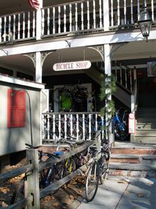 A bicycle shop with a porch, displaying bicycles on a rack outside. The entrance is marked by a sign that reads "Bicycle Shop." The shop is part of a building with a staircase leading up to the second floor, surrounded by greenery and autumn leaves.