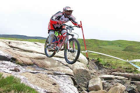 A mountain biker in a red and black outfit jumps over a rocky terrain during a competitive event, with grassy hills in the background. Fort William World Cup Downhill Track mountain bike trail.