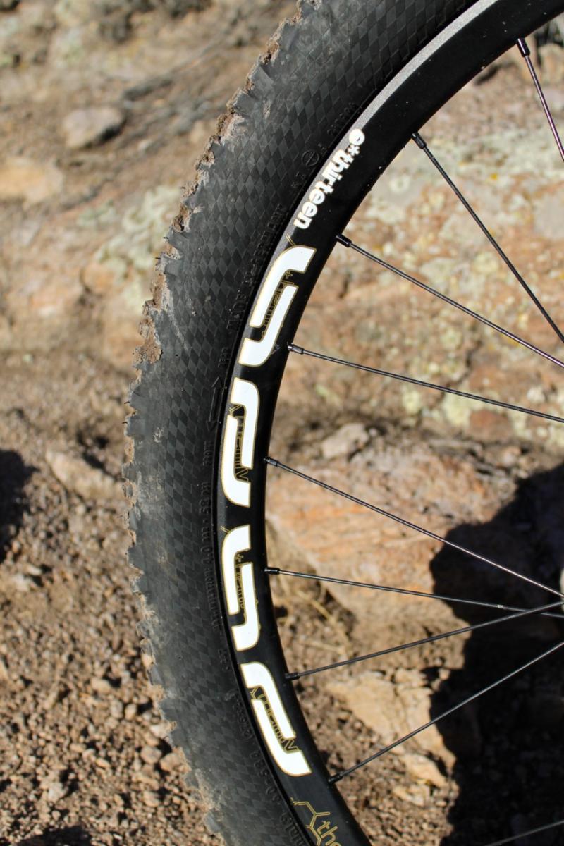 GT Force Carbon Pro: Close-up of a mountain bike tire and rim resting on rocky terrain, featuring a black tire with visible tread patterns and a decorative rim design in white. The background consists of dirt and stones, indicating an outdoor setting suitable for biking.