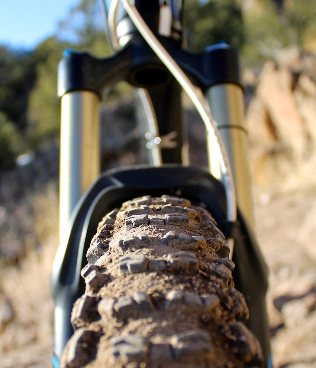 GT Force Carbon Pro: Close-up view of a mountain bike tire, showcasing the textured tread and dirt particles, with the bike's front fork and suspension visible in the background. The setting appears to be an outdoor trail with rocky terrain.