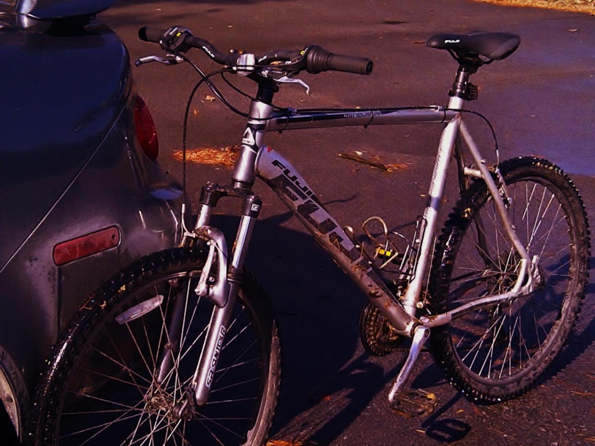 Fuji Nevada 4.0: A silver mountain bike parked next to a gray car, with a dark pavement surface and scattered leaves in the background. The bike features front and rear wheels suited for off-road riding, along with black handlebars and a comfortable seat.