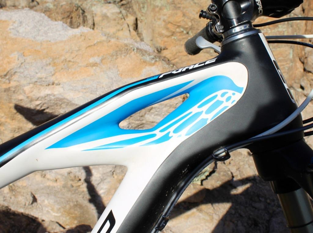 GT Force Carbon Pro: Close-up image of a bicycle frame featuring a white and blue design, with sleek lines and a prominent cutout on the top tube. The background consists of rocky terrain, highlighting the bike's outdoor setting.