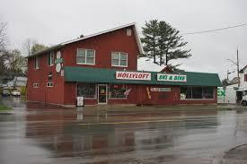 A two-story red building with a green awning displaying the sign "HOLLYLOFT SKI & DINE" on a rainy day, reflecting in the wet pavement. Pine trees are visible in the background.