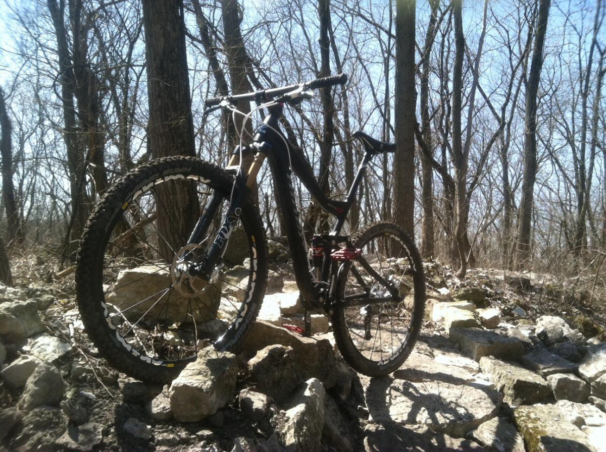 Niner W.F.O. 9: A mountain bike parked on rocky terrain in a wooded area, with bare trees in the background and a clear blue sky above. The bike has thick, treaded tires suitable for off-road riding, and it is positioned prominently among the stones.