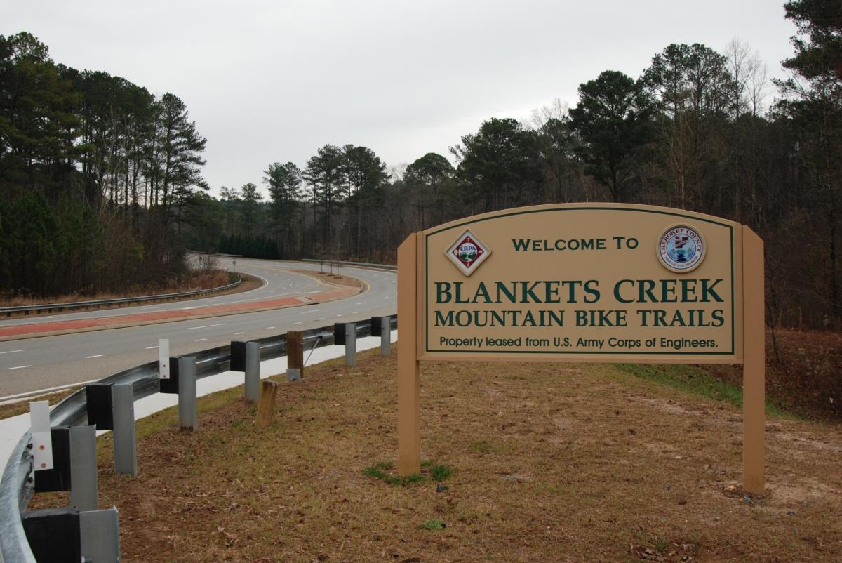 Sign welcoming visitors to Blankets Creek Mountain Bike Trails, located near a winding road, with a wooded area in the background and a cloudy sky above. The sign includes information about the trails and notes that the property is leased from the U.S. Army Corps of Engineers. Blankets Creek mountain bike trail.