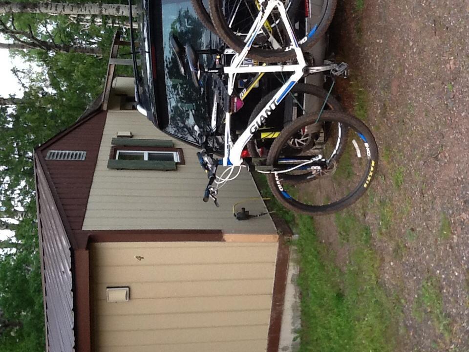 Giant Talon 29er 1: A white and blue mountain bike is mounted on the back of a black vehicle, parked in a gravel area near a cabin with a brown and beige exterior. Trees surround the setting, indicating a natural outdoor environment.