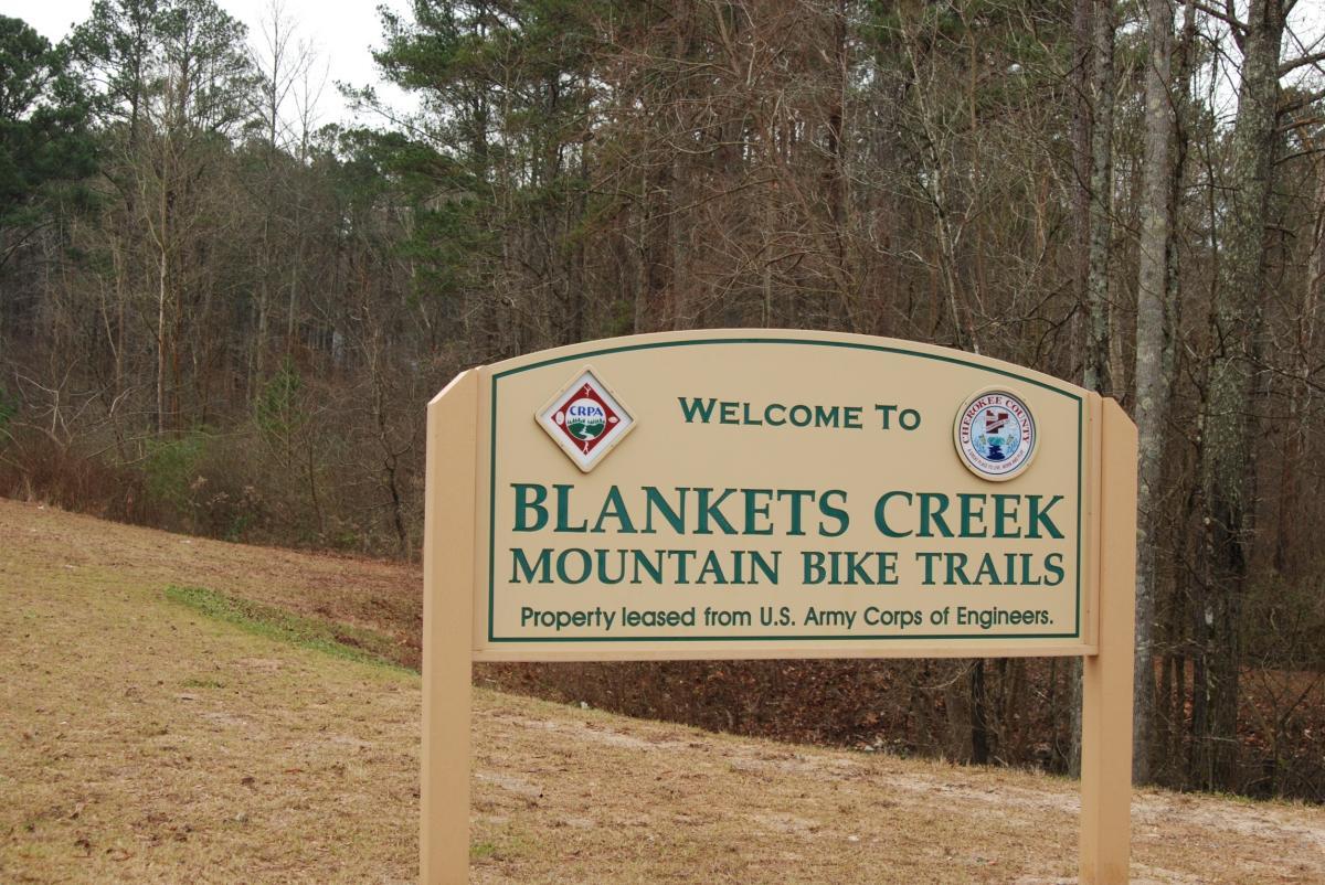 Welcome sign for Blankets Creek Mountain Bike Trails, located in a wooded area. The sign displays the name of the trails and information about property leasing from the U.S. Army Corps of Engineers. Surrounding trees indicate a natural setting for outdoor activities. Blankets Creek mountain bike trail.