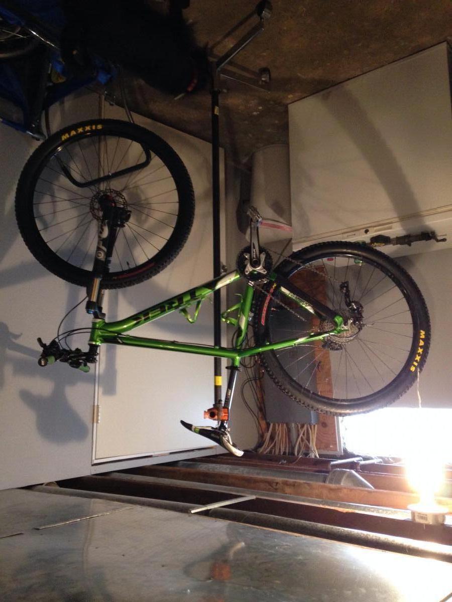 Trek Marlin: A green mountain bike is suspended upside down from a bike stand in a garage setting. The bike features thick tires with "Maxxis" branding and a pedal visible at the bottom. The background includes a dimly lit area with wires and tools, establishing a workshop atmosphere.