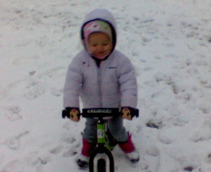 Trek Marlin: A toddler wearing a warm, puffy jacket and colorful hat is smiling while standing on a small bicycle in a snowy, outdoor setting. The ground is covered in snow, creating a winter scene.