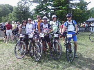 Trek Marlin: A group of six mountain bikers standing together in a grassy area, all wearing helmets and bike jerseys, with their bicycles beside them. Each cyclist has a race number displayed on their shirt. The background includes trees and a crowd of people, indicating a biking event or competition.