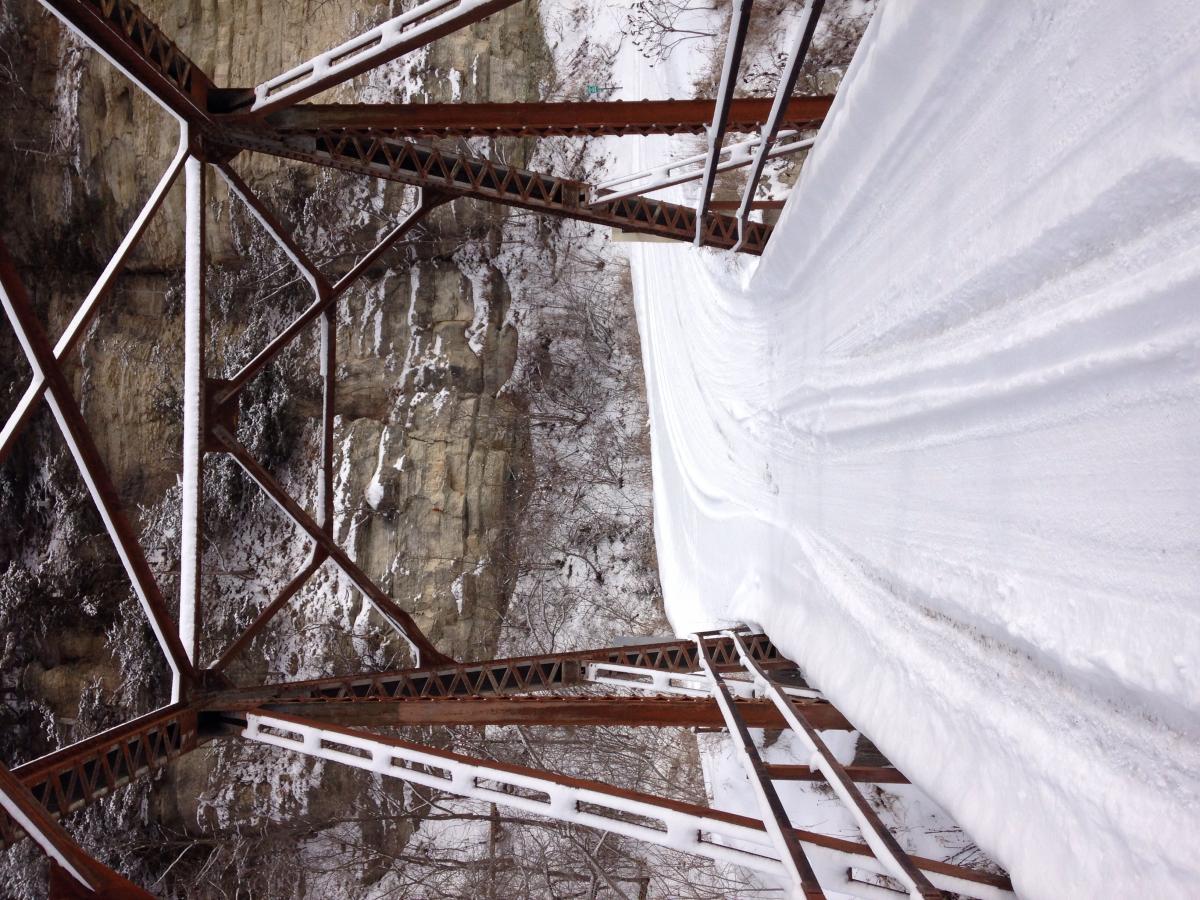 Trek Marlin: A snow-covered landscape featuring a rusted metal structure, likely a bridge or trestle, set against rocky cliffs. The foreground shows distinct tracks in the snow, indicating recent movement, while the background reveals a rugged, snowy terrain.