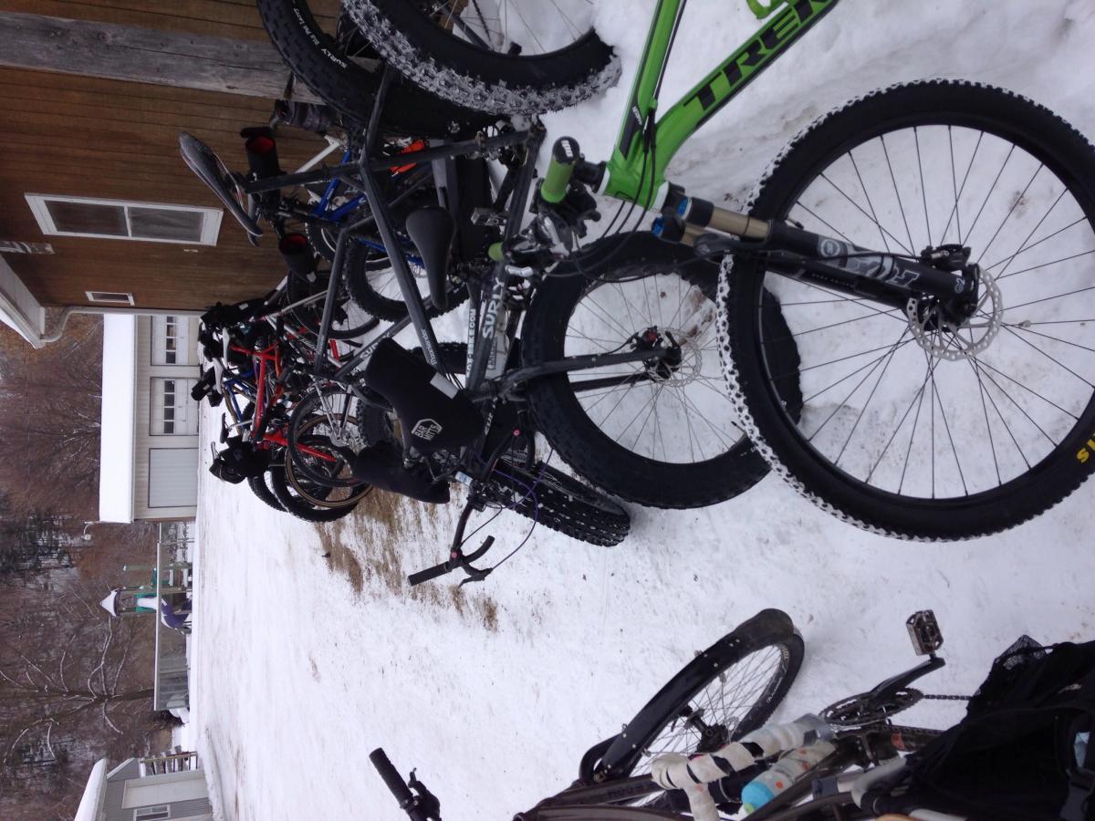 Trek Marlin: A group of parked bicycles on snow-covered ground, with a wooden building in the background. The bikes are a mix of colors and styles, including mountain bikes with thick tires designed for winter riding. Snow blankets the surrounding area, and a playground with a slide can be seen in the distance.