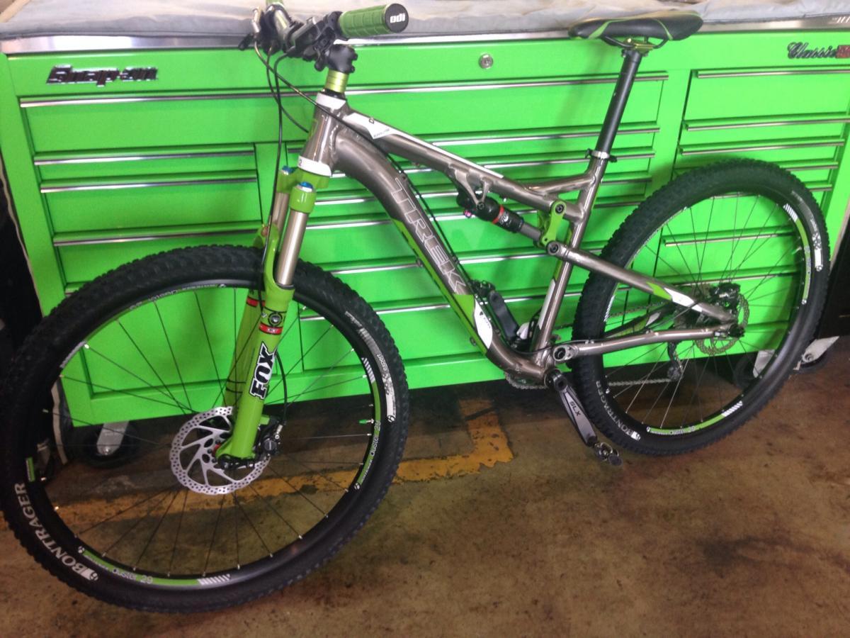 Trek Rumblefish Elite: A mountain bike with a sleek aluminum frame featuring a combination of silver and green components, positioned in front of a bright green tool chest. The bike has thick tires suitable for off-road riding and is equipped with a Fox suspension fork.