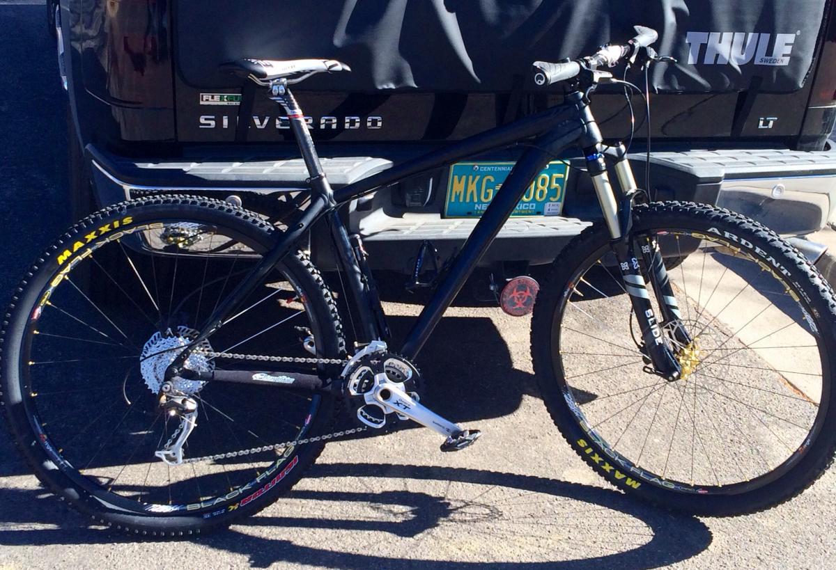 Specialized Rockhopper Comp 29: A black mountain bike parked beside a pickup truck. The bike features thick tires, a disc brake system, and a sleek frame design. The background shows the tailgate of a silver pickup truck, with a Thule bike rack visible, alongside a New Mexico license plate.