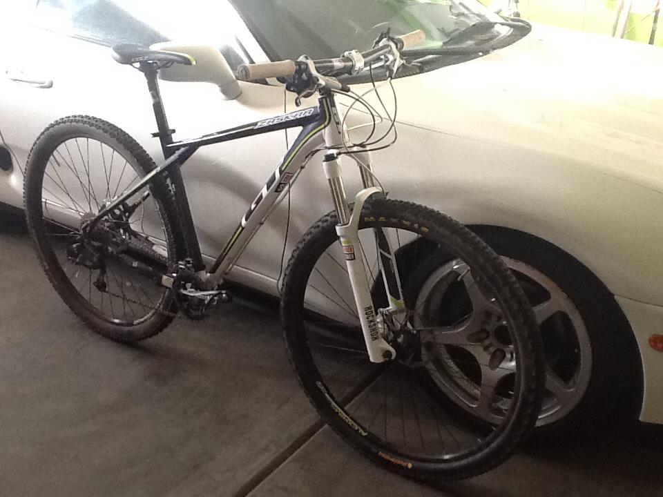 GT Zaskar 9r Comp: Mountain bike leaning against a white car, showcasing its black and silver frame, thick tires, and front suspension fork. The setting appears to be a garage with a neutral floor and part of the car visible in the background.