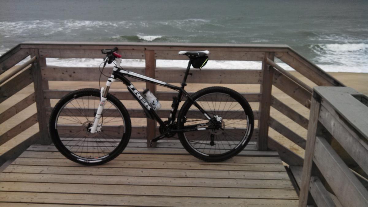 Trek X-Caliber 8: A black and white mountain bike parked on a wooden deck overlooking the beach, with waves in the background and a cloudy sky.