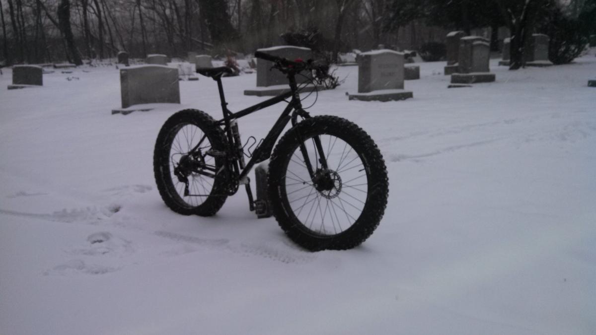 Trek X-Caliber 8: A black fat tire bike standing on snow-covered ground in a cemetery. Snow falls gently, and gravestones are visible in the background, creating a serene, wintry scene.