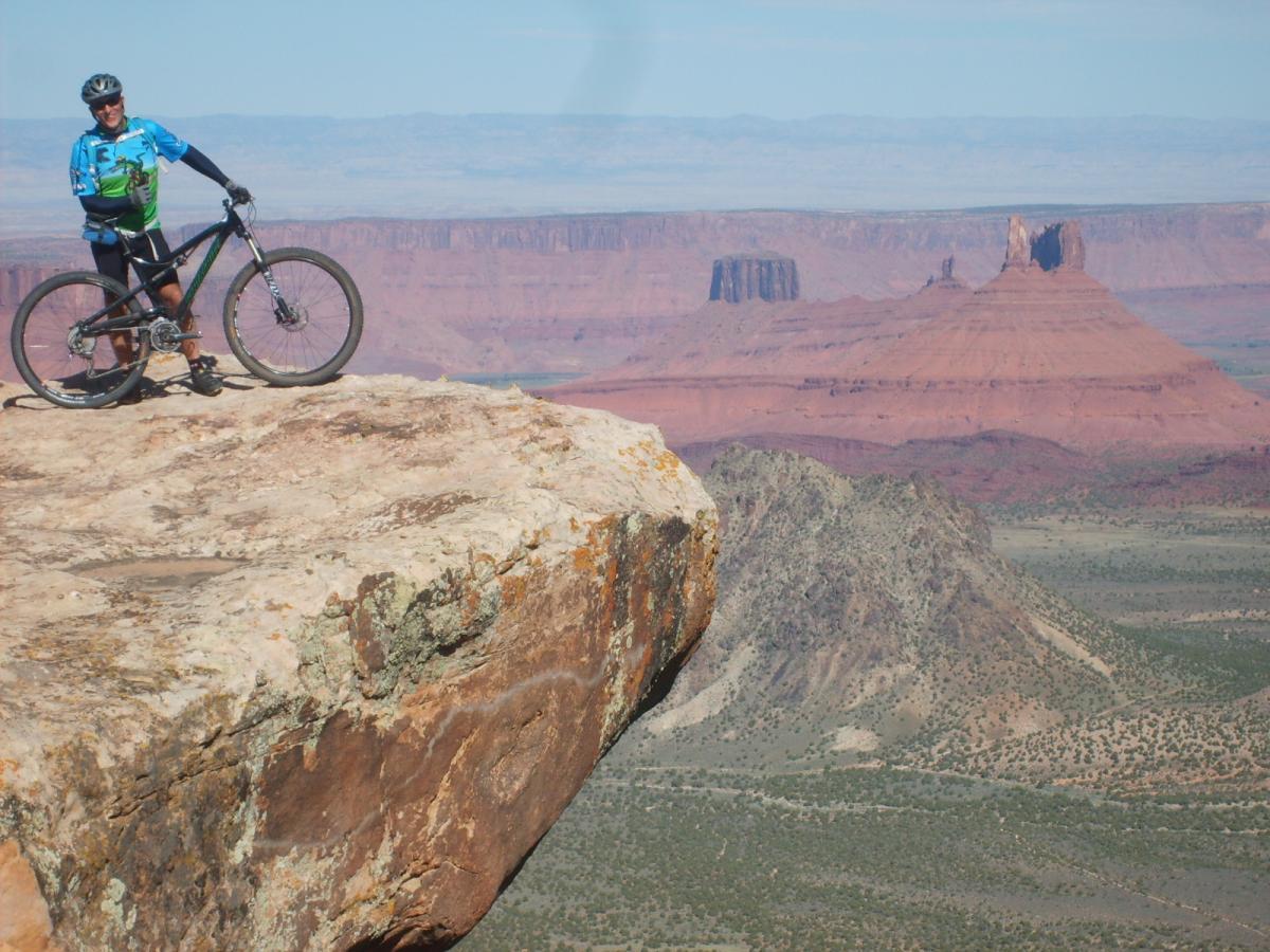Giant Trance X2: A mountain biker in a blue and green jersey stands confidently on a large rock outcrop, overlooking a vast desert landscape with mesas and red rock formations in the background. The sky is clear, and the scene captures a sense of adventure and the beauty of nature.