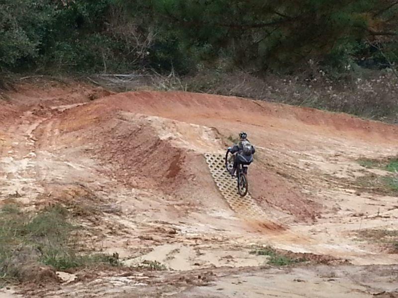 A person riding a mountain bike on a dirt track with mounds and a textured ramp in a natural outdoor setting. The terrain is predominantly reddish-brown dirt with patches of grass and vegetation in the background. Tom Brown / Lafayette Heritage Park mountain bike trail.