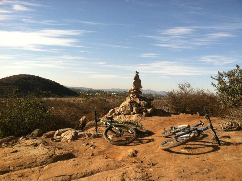 Two bicycles are resting on a rocky, dirt trail overlooking a scenic landscape. In the background, there is a stone cairn stacked neatly, with rolling hills and a clear blue sky above. The area is surrounded by sparse vegetation and the distant skyline adds depth to the peaceful outdoor setting. Lake Calavera mountain bike trail.