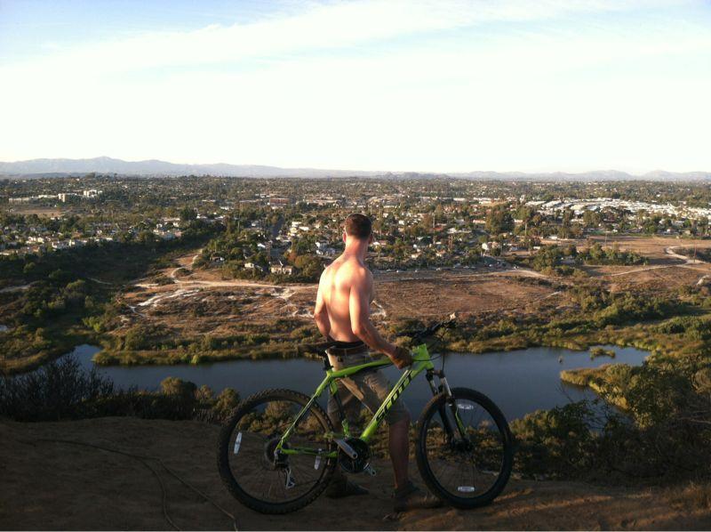 A shirtless person stands next to a mountain bike, looking out over a scenic landscape that includes a river, fields, and distant buildings under a clear blue sky. The sun is setting, casting a warm glow over the scene. Lake Calavera mountain bike trail.