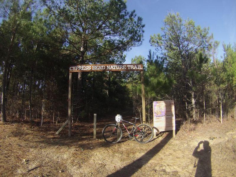 A mountain bike parked beside the entrance sign for the Cypress Bend Nature Trail, surrounded by tall pine trees and sunlight filtering through the foliage. The trailhead features a wooden sign with the trail name and additional information nearby. Cypress Bend Resort mountain bike trail.