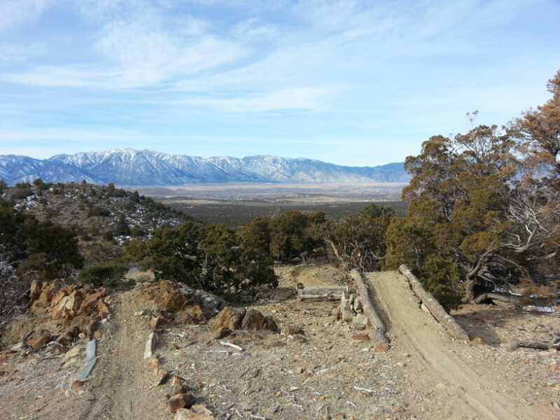A scenic view of a mountainous landscape with snow-capped peaks in the background, surrounded by rolling hills and sparse vegetation. A dirt path leads through the foreground, with rough terrain and scattered rocks, while green shrubs dot the landscape. The sky is bright and clear, creating a serene outdoor atmosphere. Pine Nut mountain bike trail.