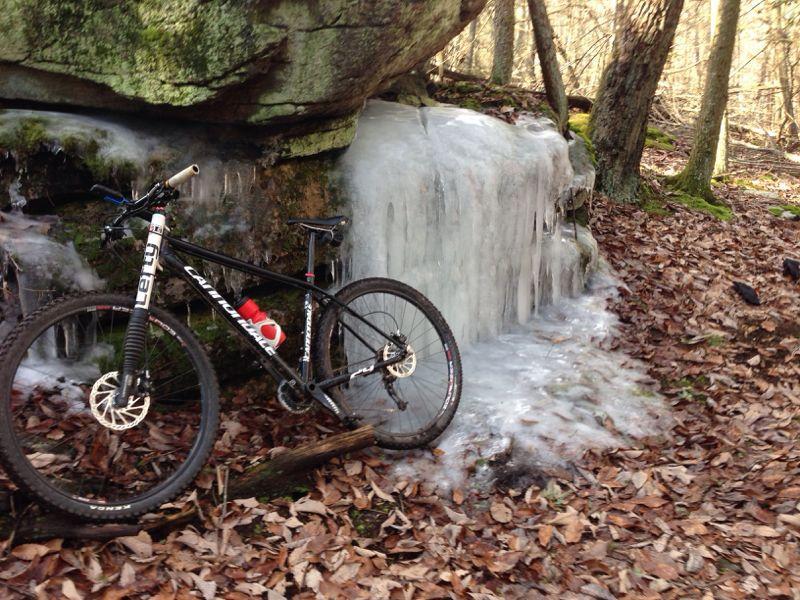 A black mountain bike rests beside a frozen waterfall in a forest, with large rocks and trees surrounding it. The ground is covered in brown leaves, and the sunlight filters through the trees, illuminating the icy formation. Frederick Watershed mountain bike trail.