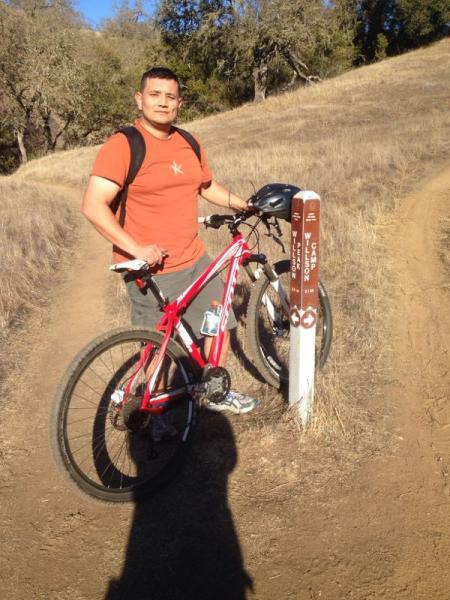A person standing next to a mountain bike on a dirt path, with a trail marker indicating the direction. The rider is wearing an orange shirt and shorts, with a backpack. The background features tall grass and trees under a clear blue sky. Henry W. Coe State Park mountain bike trail.